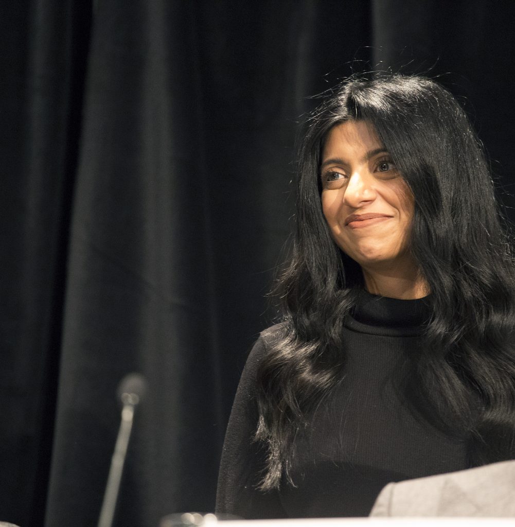 A woman standing at a podium in front of a black curtain, smiling.