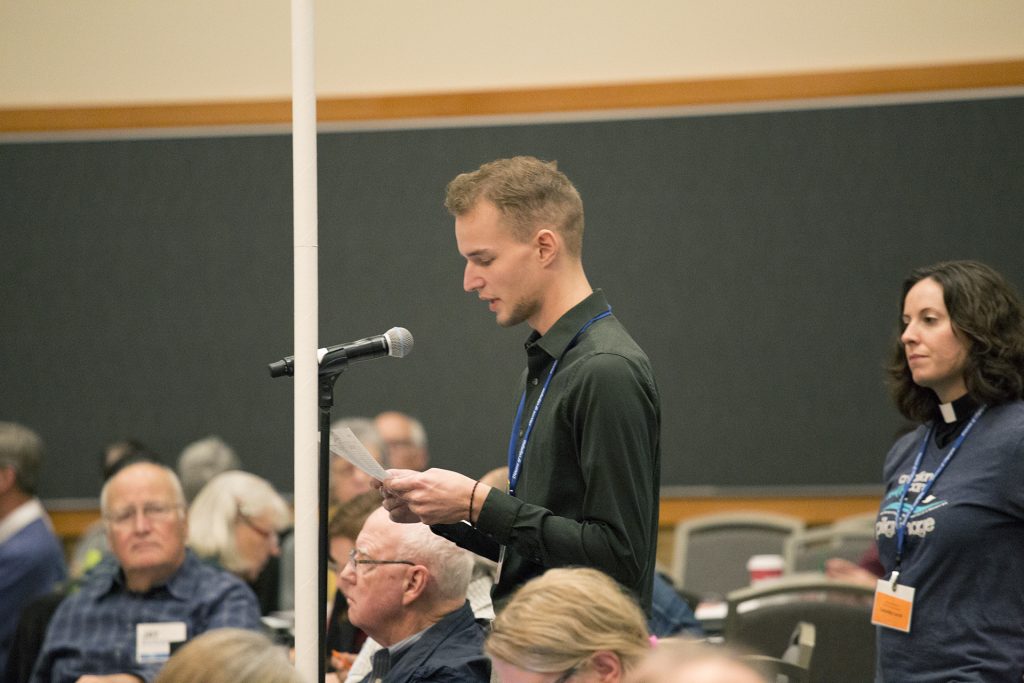 A young man stands at a microphone, reading from a paper in a room with people sitting and a female priest standing behind him